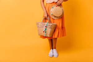 Garota bronzeada em saia laranja e sapatos brancos em pé segurando bolsa de palha decorada com pompons e na outra mão um chapéu