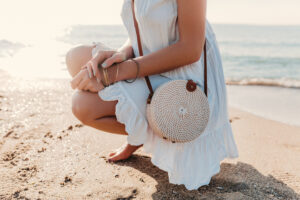 Modelo agachada na areia, segurando uma bolsa de palha para casamento na praia.